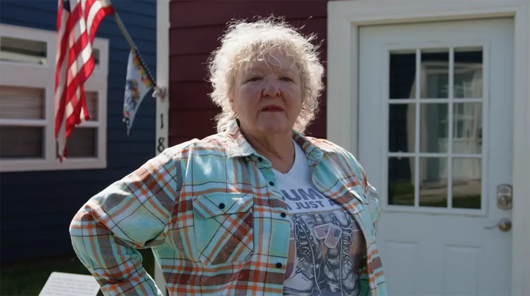 a veteran in close-up, standing in front of a house and an American flag